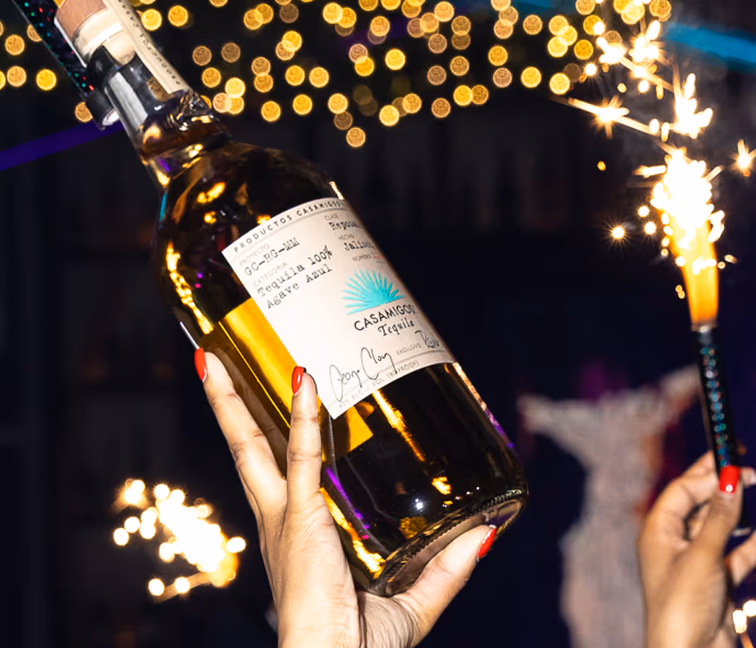 Hand with red nail polish holding a bottle of Casamigos tequila with sparklers in the background and festive lights.