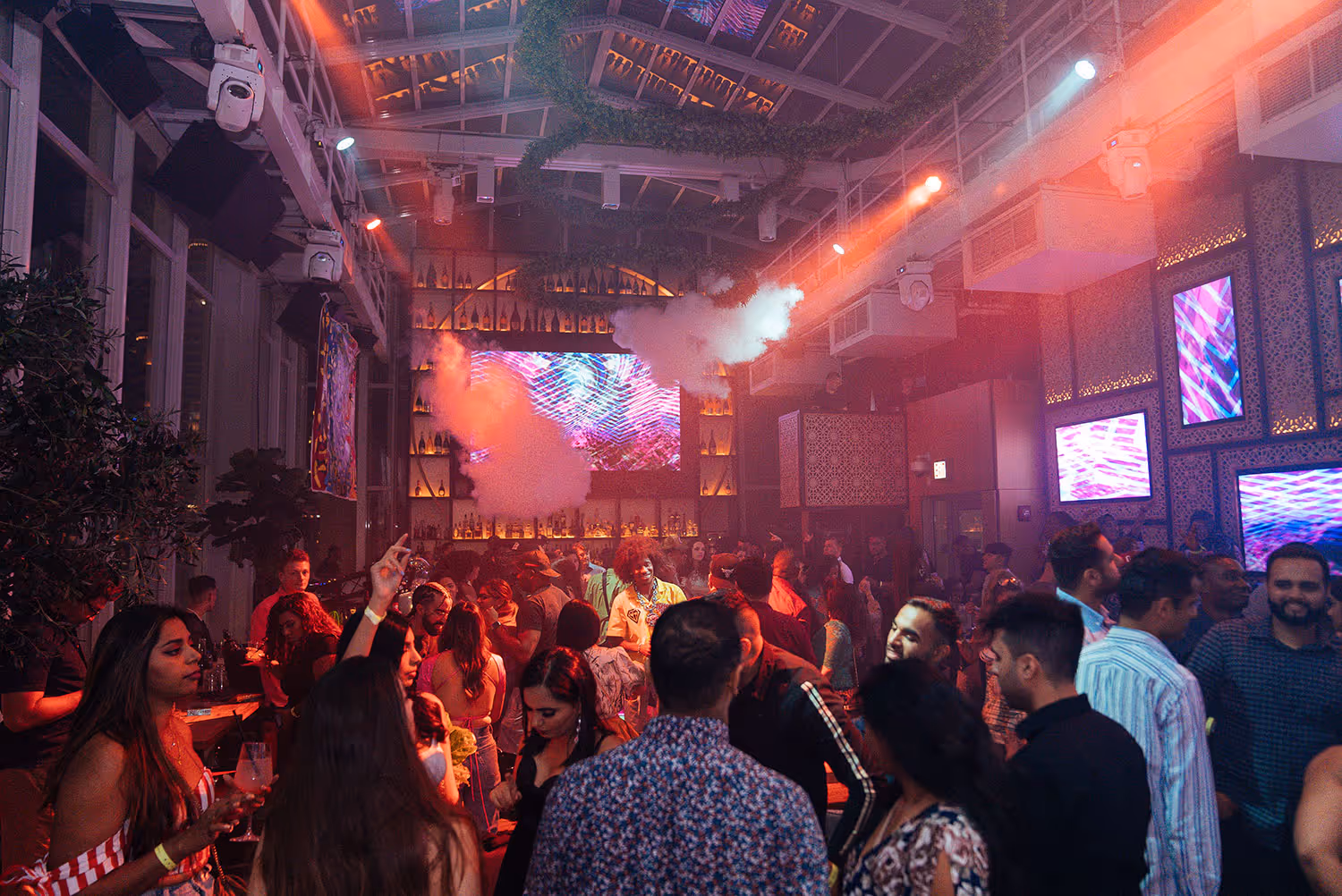 Crowded nightclub interior with people dancing, colorful lights, smoke effects, and digital screens on the walls.