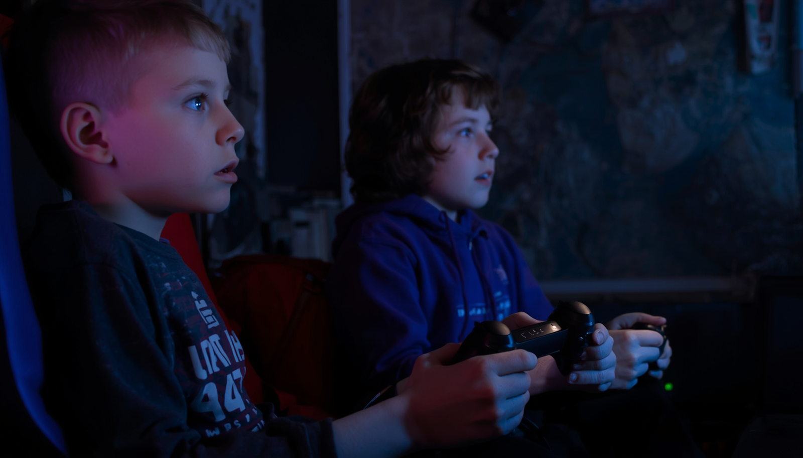 Two boys sitting side by side playing video games with focused expressions in a dimly lit room.