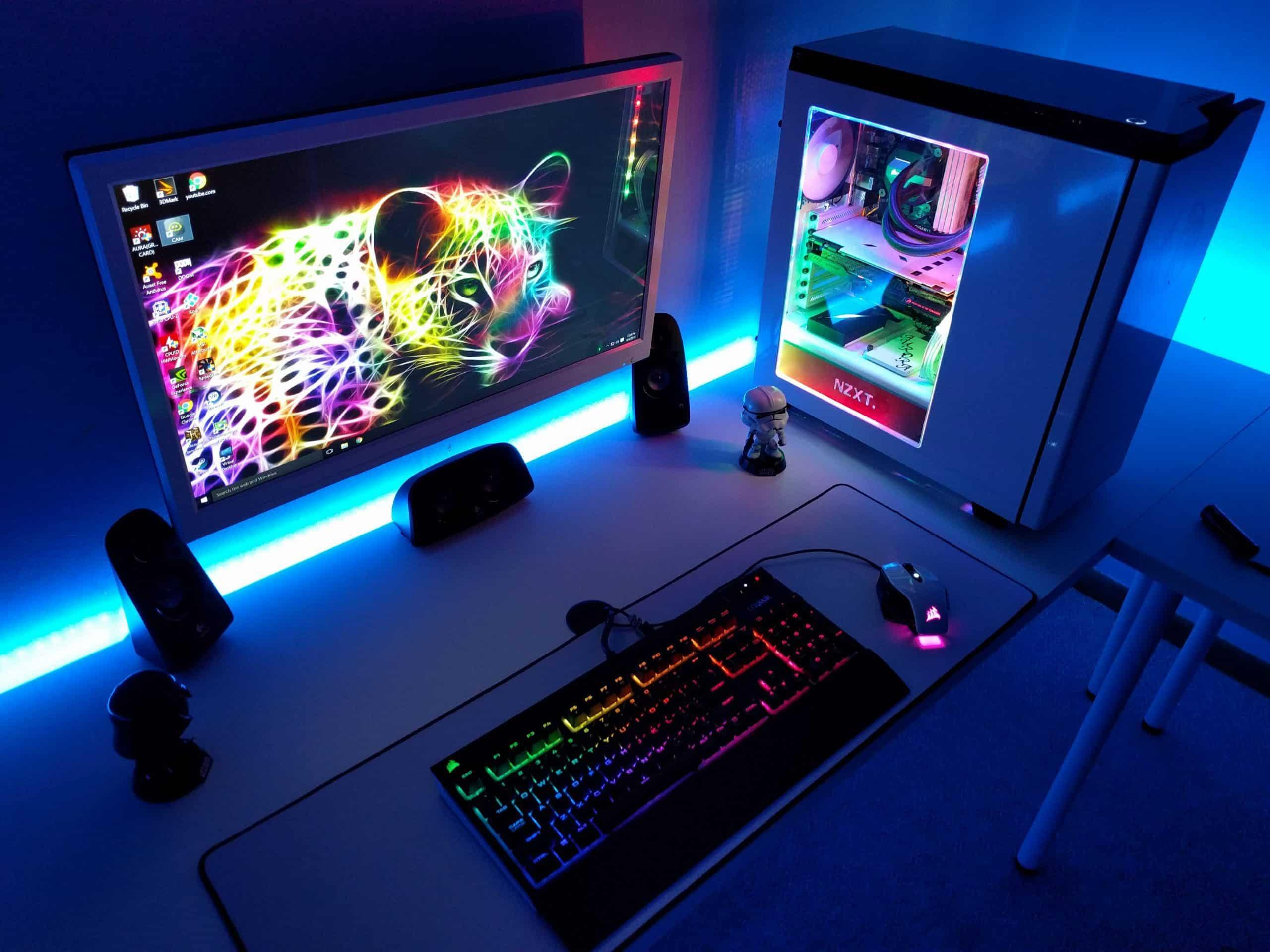 Gamer desktop setup with a monitor displaying a neon-colored leopard wallpaper, RGB backlit keyboard and mouse, and a white gaming PC with visible internal components and RGB lighting.