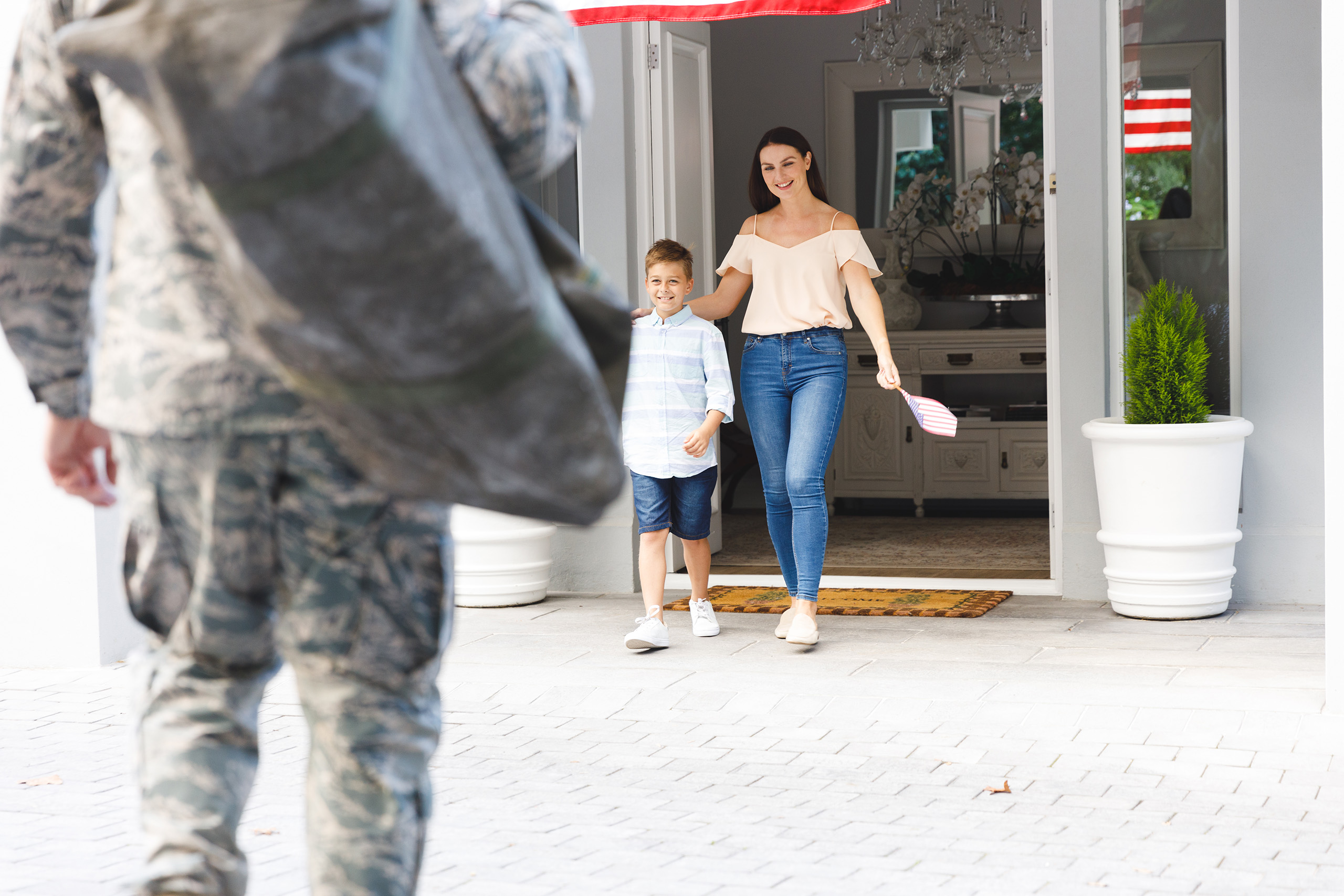 A military person approaches their home. A woman and a child holding a small American flag stand at the door, smiling and appearing pleased.