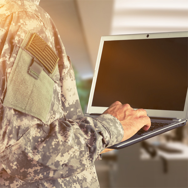 A person in military camouflage holding a laptop, emphasizing technology in defense. The setting is bright and indoors, suggesting a secure environment.