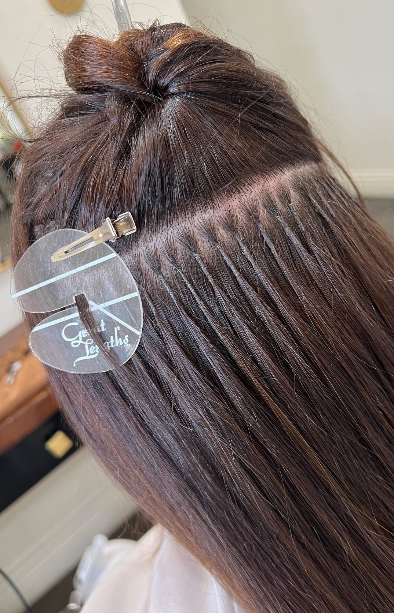Close-up of a woman’s hair during a professional hair extension installation. The stylist has sectioned the top layer of brown hair and clipped it up with a transparent “Great Lengths” branded clip. A neat row of small, evenly spaced bonded hair extensions is visible near the scalp, showing precise application technique on smooth, shiny natural hair.