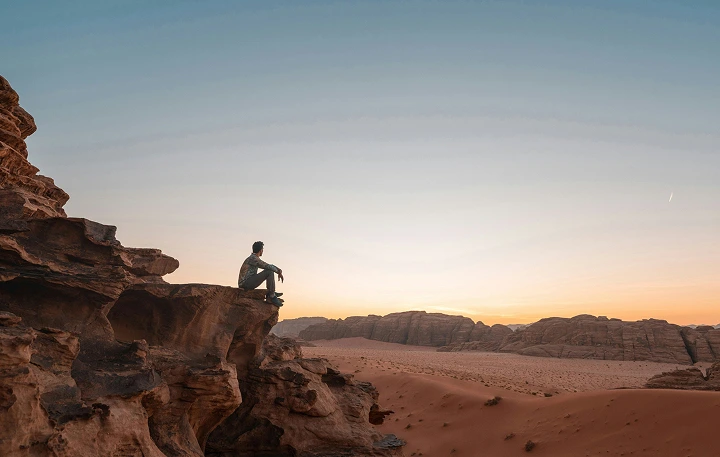 Person sited alone in a rock located in a mountain with a sunny sky behind