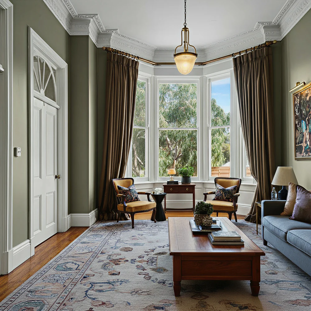 Bright, elegant living room with bay windows, a wooden coffee table, and comfortable seating, featuring decorative curtains and plants.