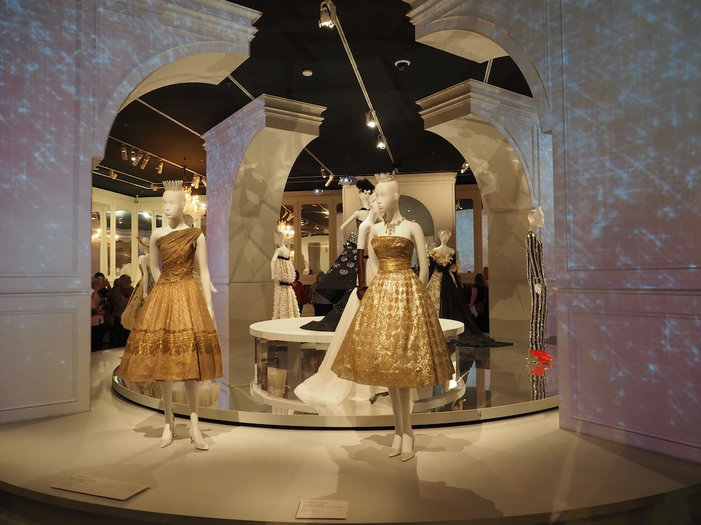An NGV exhibit featuring elegant dresses displayed on mannequins in a stylish gallery setting. The decor includes arches and ambient lighting.