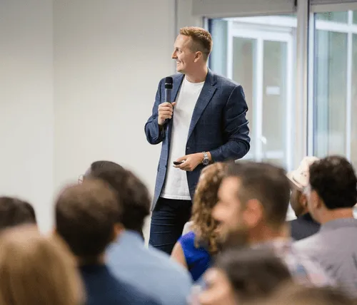 Man in blue suit speaking with microphone to audience in conference room, with large windows in background.