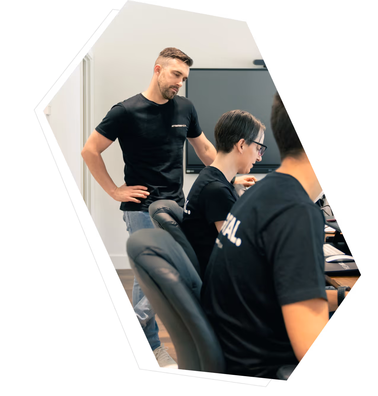 Three people in black T-shirts working together in an office, one standing and two seated at desks with computers.