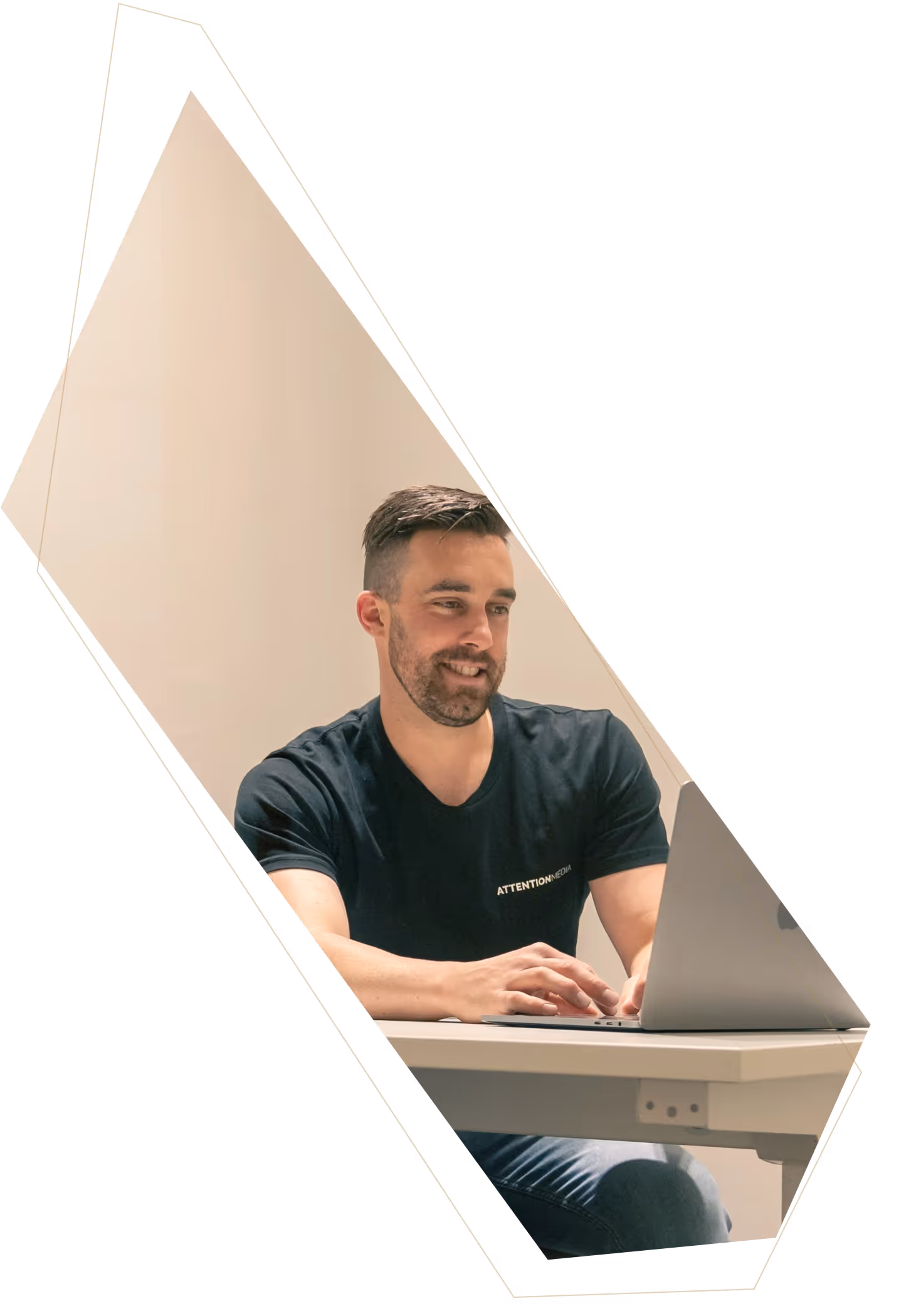Man with short dark hair and beard wearing a black T-shirt sitting at a white desk, smiling and typing on a laptop.