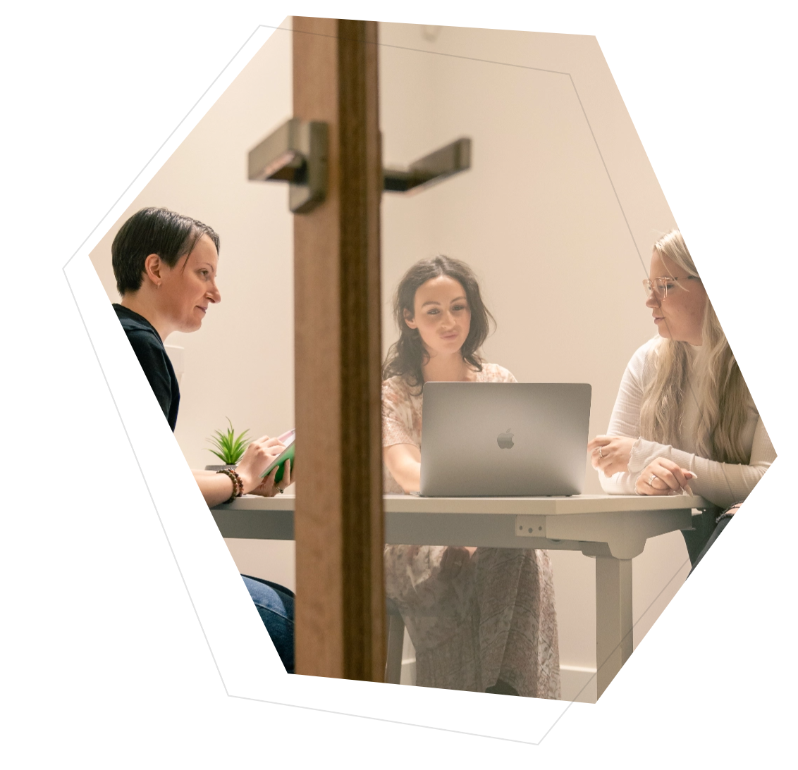 Three women sitting around a table in a meeting room, one using a laptop while the others listen and take notes.