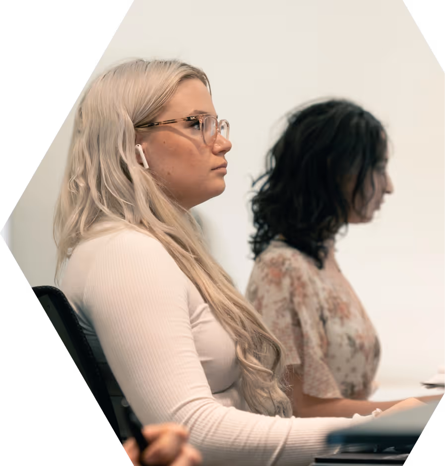 Side profile of a woman with long blonde hair and glasses wearing a white top, focused and sitting in an office setting with another woman working in the background.