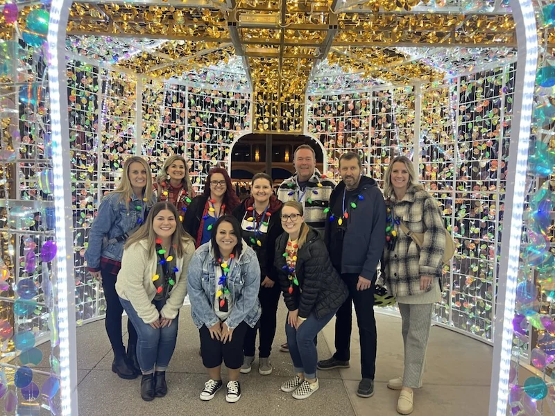 Group of ten people smiling and posing inside a brightly lit festive structure decorated with multicolored lights and gold ornaments.