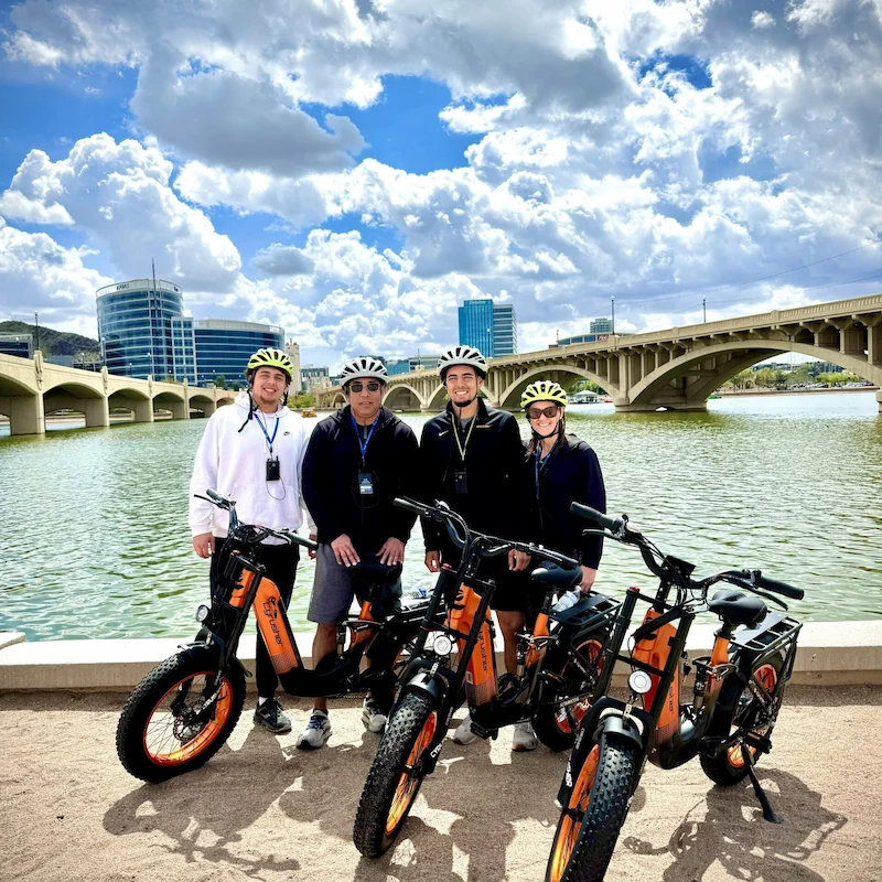 Four people wearing helmets standing with electric bikes on a riverside path with a bridge and city buildings in the background under a partly cloudy sky.