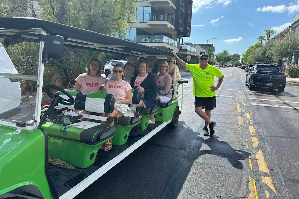Group of people sitting in a green open-air shuttle vehicle on a sunny urban street, with a man in a bright yellow shirt standing beside it.