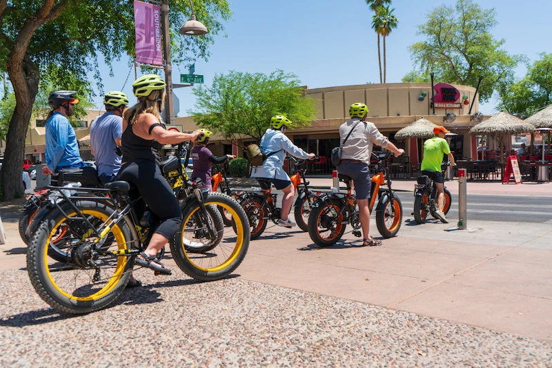 Group of people wearing helmets riding electric fat-tire bikes on a sunny urban street near shops and palm trees.