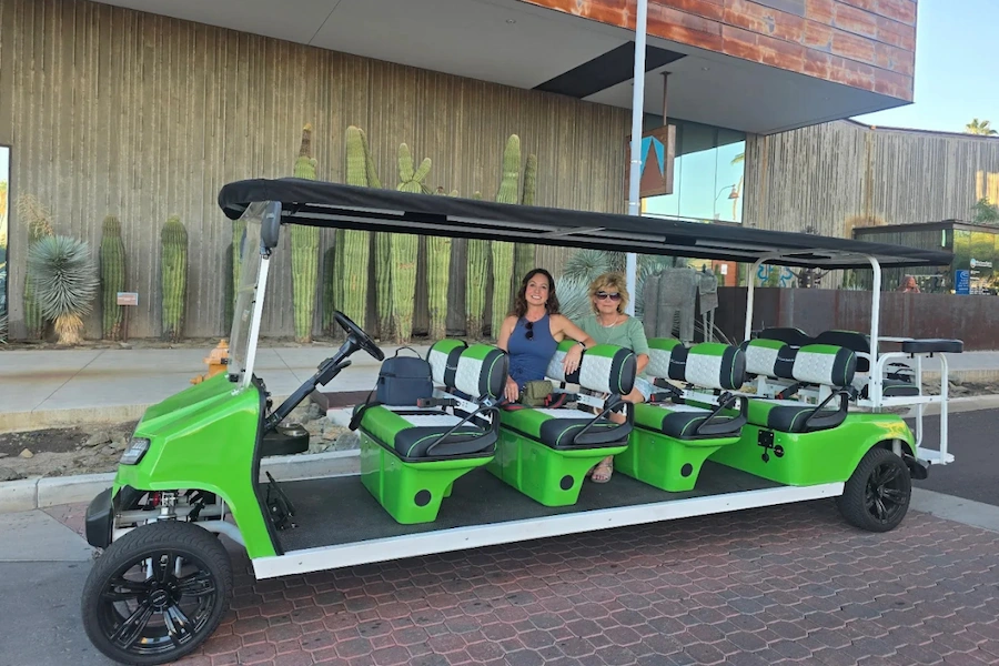 Two women sitting on the middle seats of a green open-air golf cart parked on a street with cacti and wooden buildings in the background.