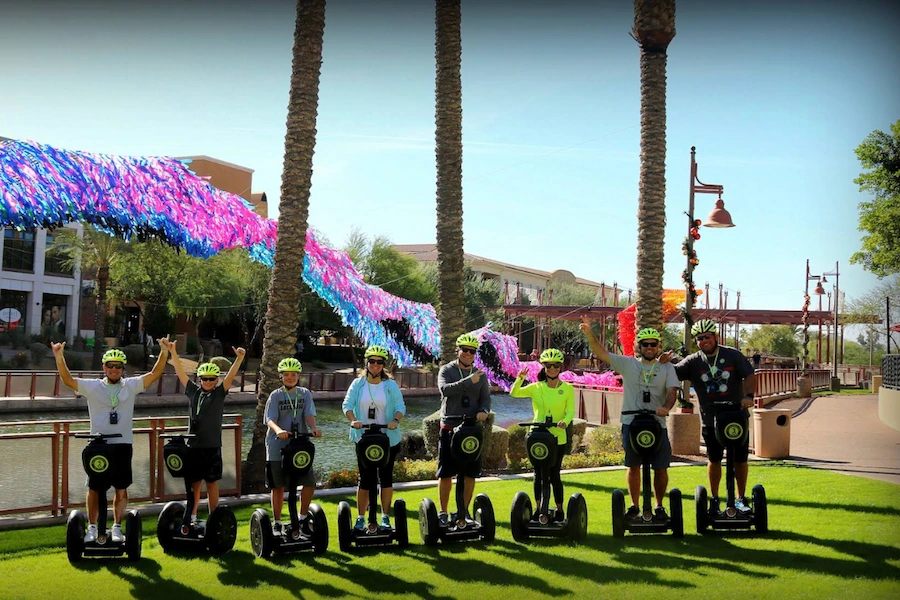 Group of eight people wearing helmets and riding Segways on green grass beside a canal with colorful decorations overhead.