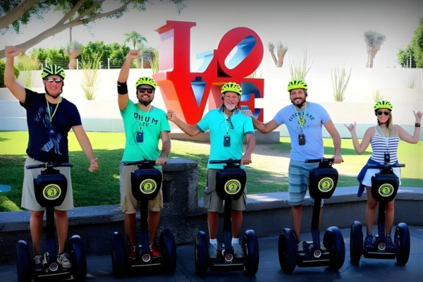 Five people wearing helmets standing on Segways, smiling and raising their arms in front of a large red and blue LOVE sculpture outdoors.