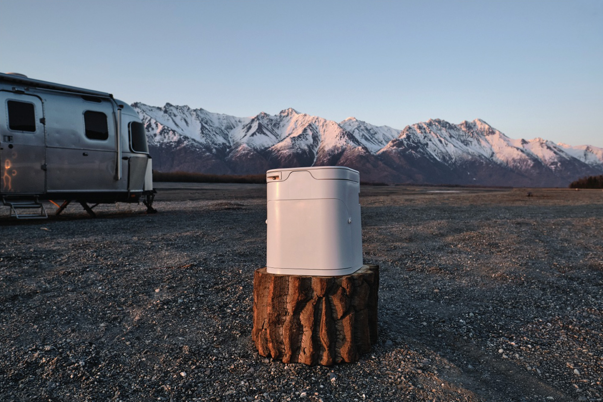 OGO composting toilet on a tree stump beside a silver travel trailer, with snow-capped mountains behind