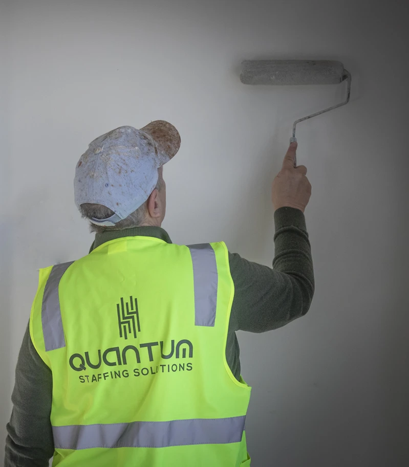 A worker in a Quantum Staffing Solutions hi-vis vest painting a wall with white paint.