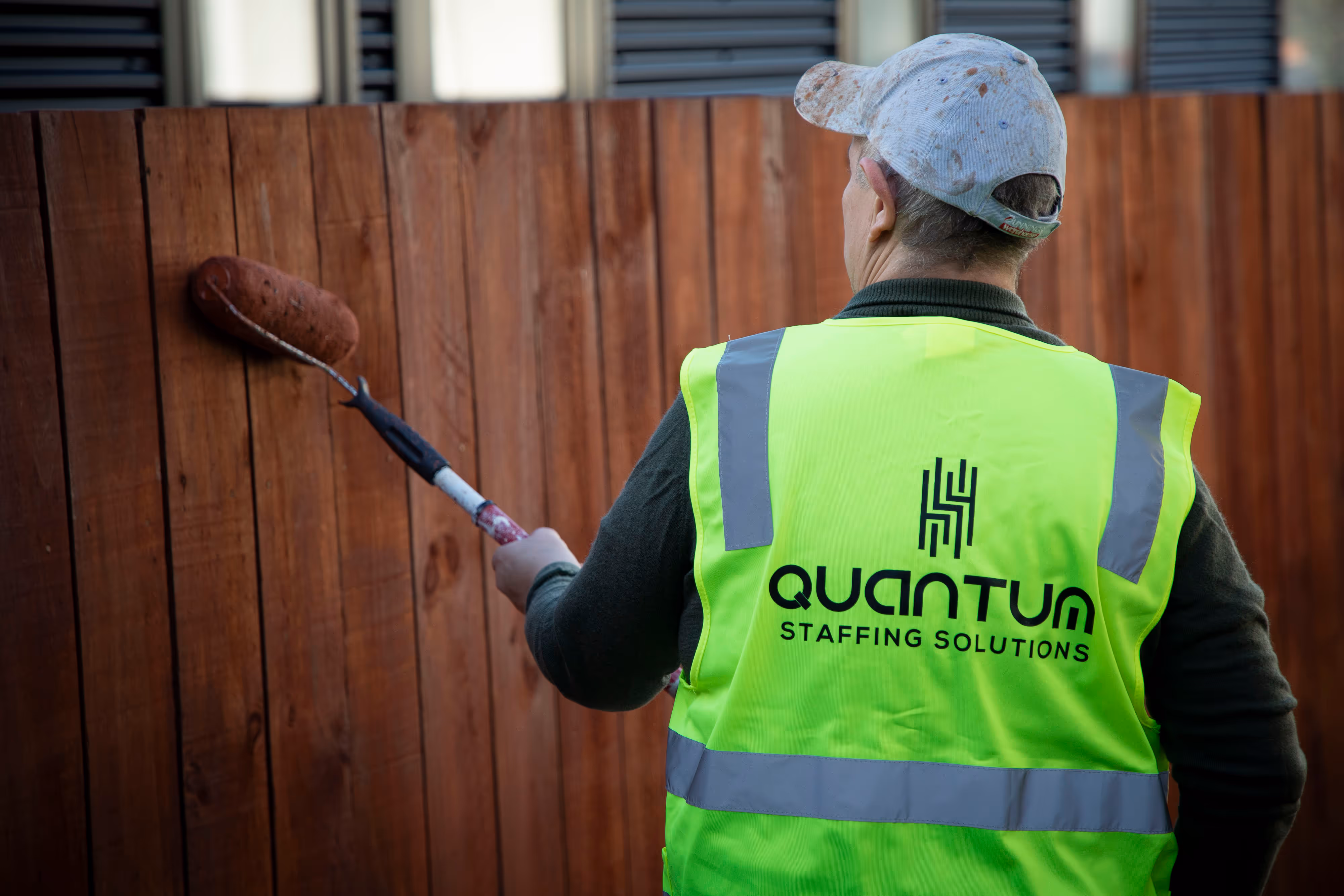 A worked wearing a Quantum Staffing Solutions branded hi-vis vest painting a fence.