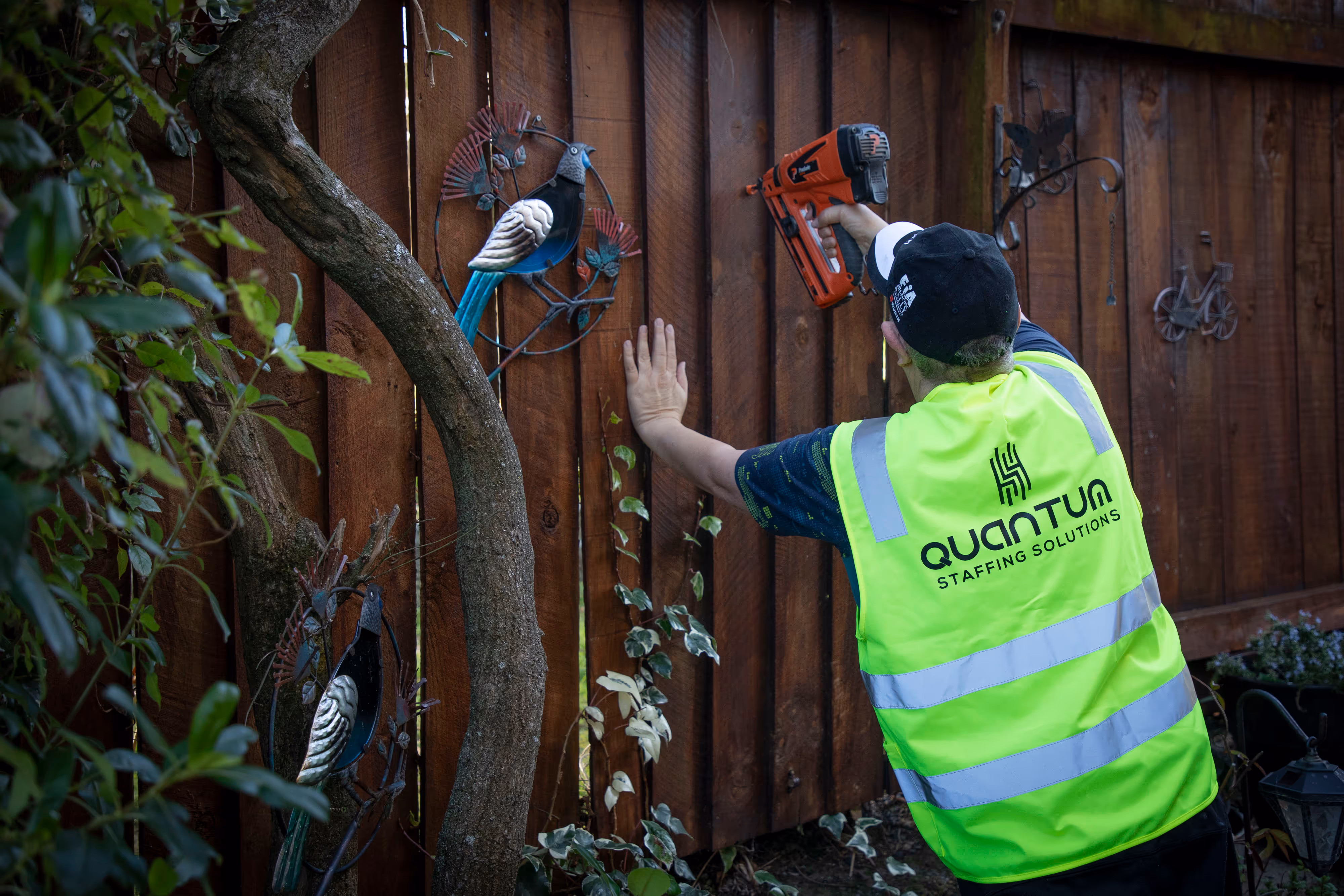 A worker in a hi-vis vest holding a nail gun against a wooden fence outside.