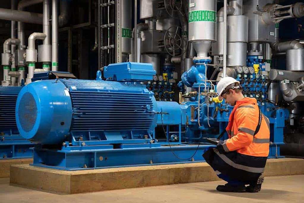 Worker in orange safety jacket and white helmet inspecting a large blue industrial machine in a factory setting.
