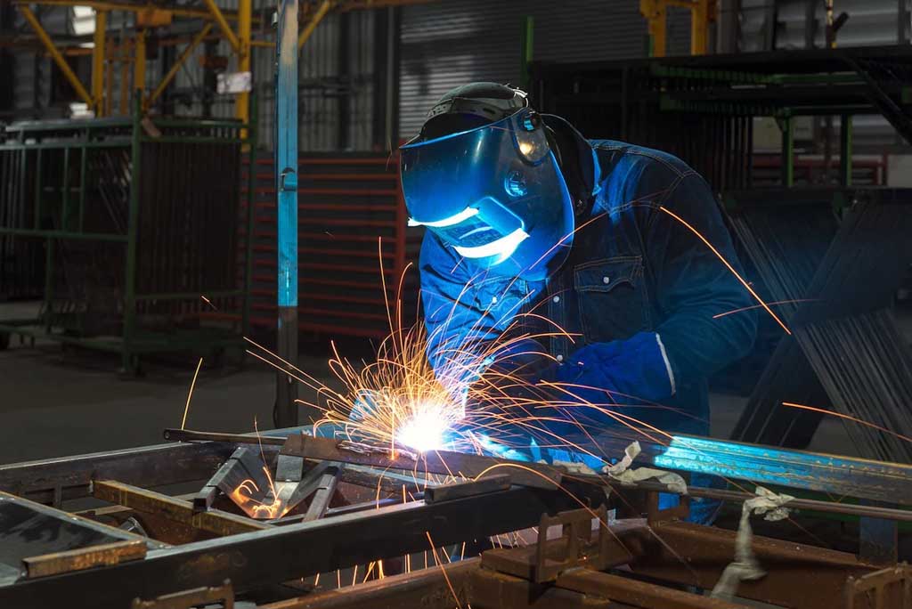 Worker wearing a protective welding helmet and gloves welding metal with sparks flying in an industrial workshop.