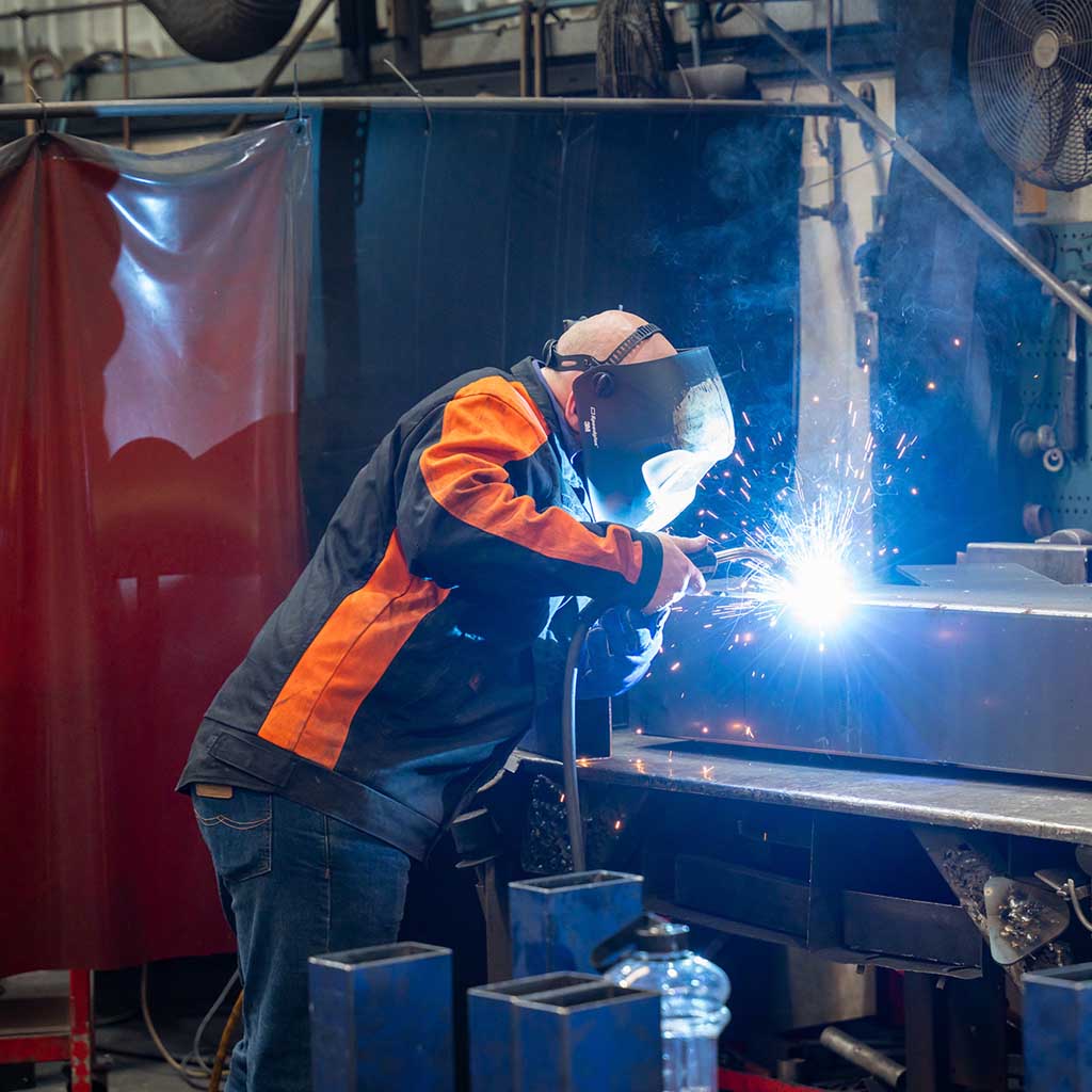 Worker wearing protective welding helmet and jacket welding metal, sparks flying in workshop.