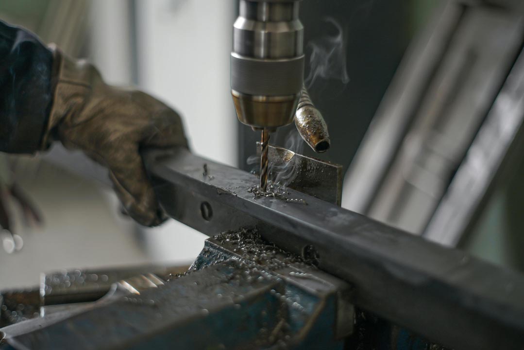 Gloved hand operating a drill press to bore holes in a metal bar with metal shavings around.