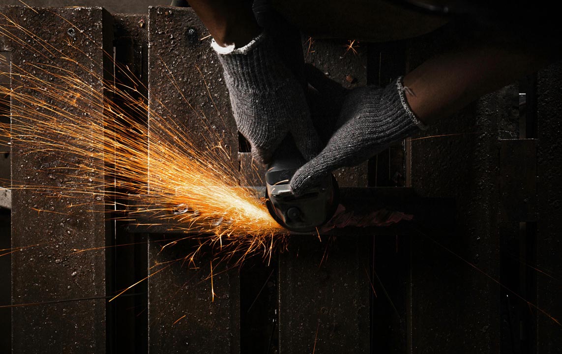 Hands wearing gray gloves using a grinder tool emitting bright orange sparks while working on metal pieces.