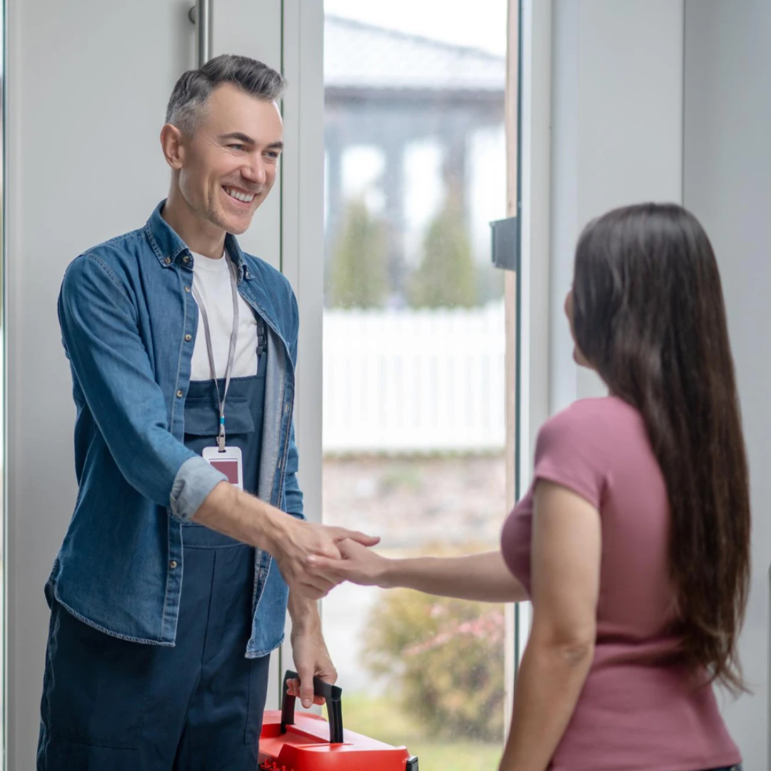 Smiling male technician shaking hands with a woman at her doorstep while holding a red toolbox.