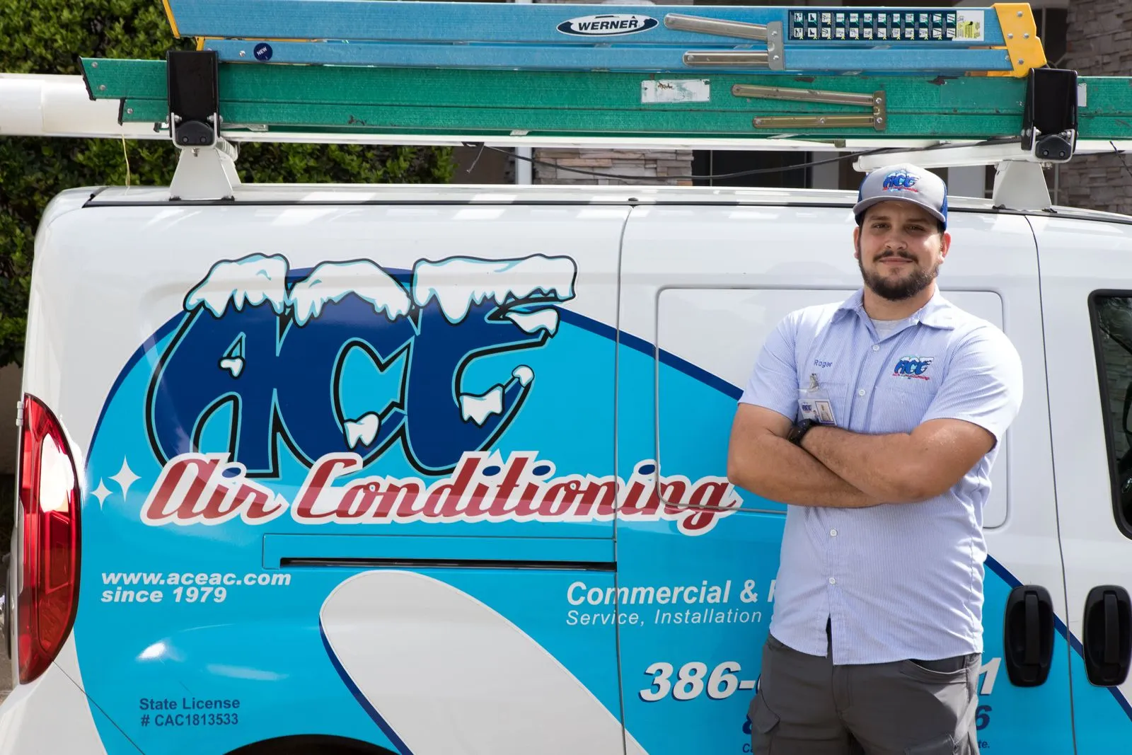 Technician standing with arms crossed next to a white and blue Ace Air Conditioning service van with ladders on top.
