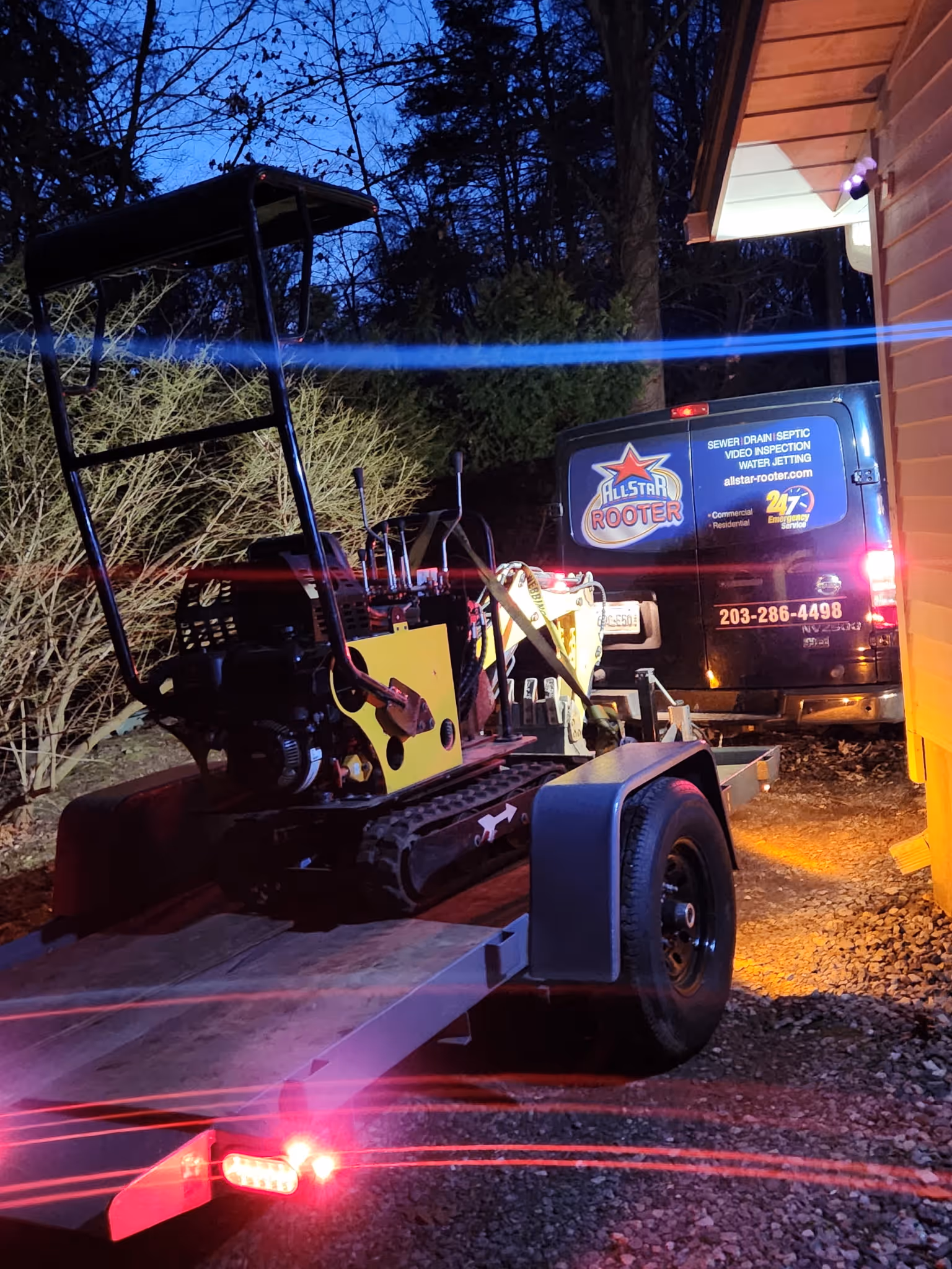 Small excavator loaded on a trailer attached to an Allstar Rooter service van parked beside a house at dusk.