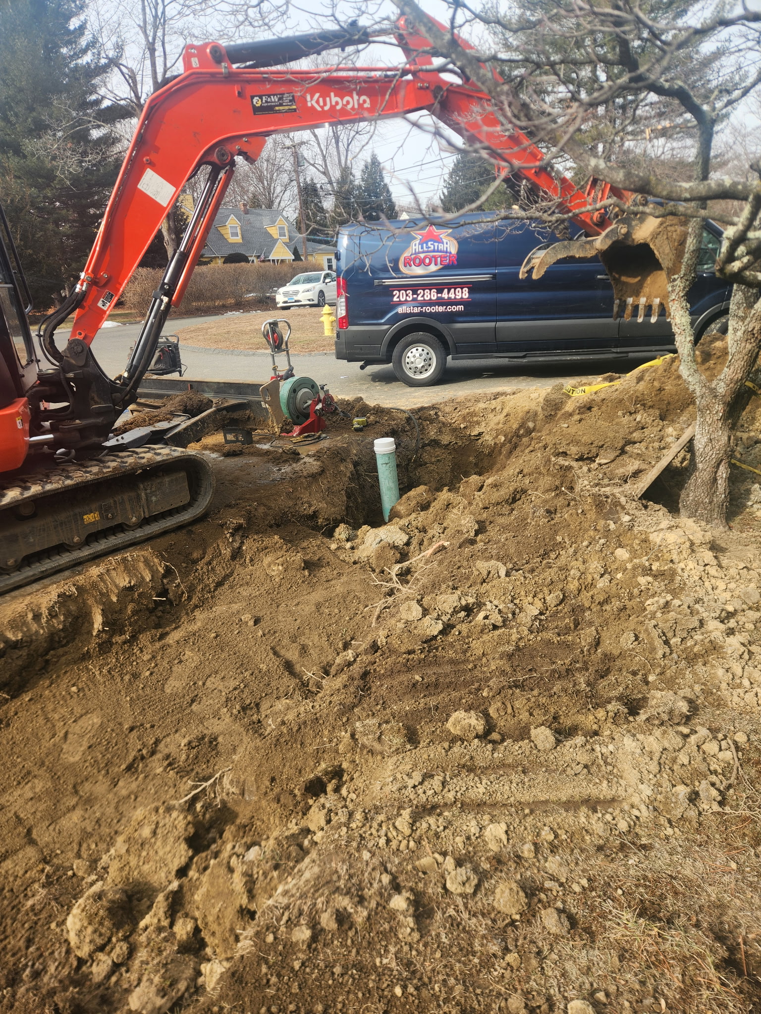Excavator digging a large hole in front of a tree with a plumbing pipe exposed and an All Star Rooter van parked on the street.