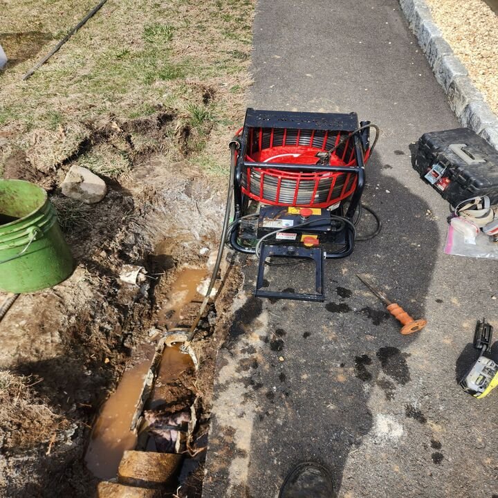 Sewer cleaning equipment including a drain snake machine and tools beside a muddy trench with exposed pipes.