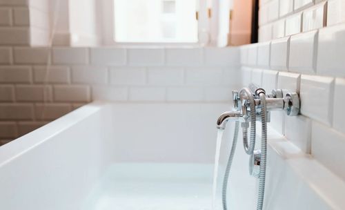 Modern white bathtub with water flowing from a chrome faucet against a white tiled wall.