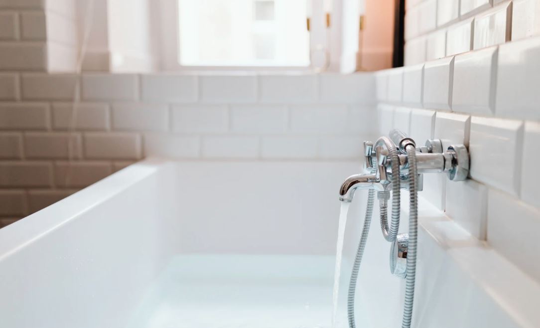 Modern white bathtub with water flowing from a chrome faucet against a white tiled wall.
