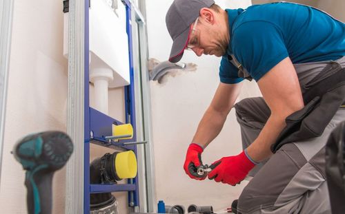 Worker wearing red gloves and safety glasses installing plumbing fixtures in a bathroom under construction.
