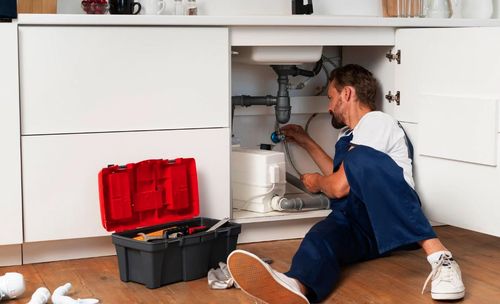Plumber in blue overalls working on pipes under a kitchen sink with an open toolbox nearby.