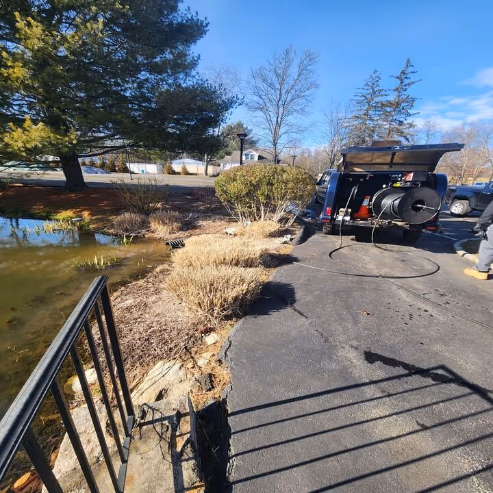 Asphalt pavement separating a pond from bushes with a truck parked on the right side, equipped with a sewer cleaning hose.