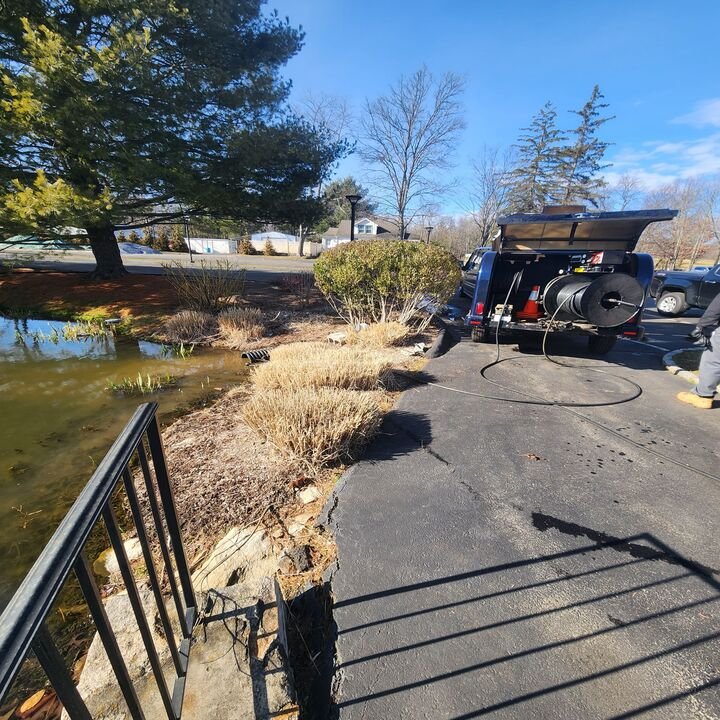 Asphalt pavement separating a pond from bushes with a truck parked on the right side, equipped with a sewer cleaning hose.