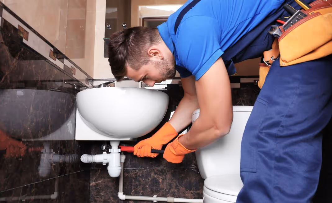 Plumber in blue uniform and orange gloves fixing a pipe under a bathroom sink.