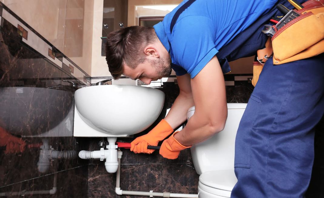 Plumber in blue uniform and orange gloves fixing a pipe under a bathroom sink.