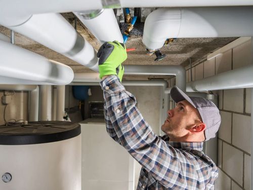 Plumber in a plaid shirt and gray cap adjusting white pipes against a concrete wall.
