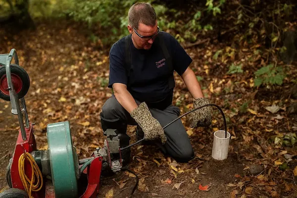 Man wearing gloves and safety glasses operating a drain cleaning machine outdoors surrounded by fallen leaves.