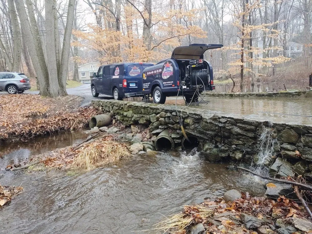 Service van with Allstar Rooter logo parked on a stone bridge over a small creek with water flowing through pipes beneath.