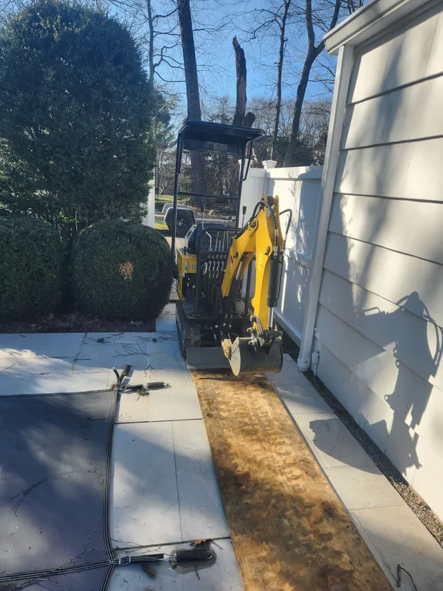 Yellow mini excavator parked on a wooden plank beside a white fence and a house, next to trimmed bushes.