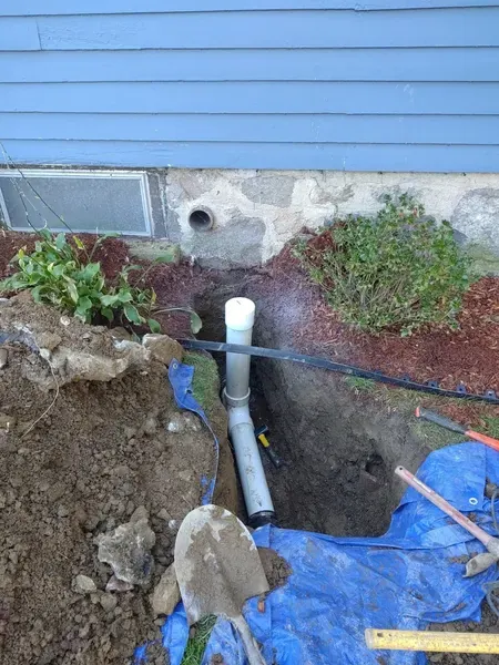 Trench beside a blue house with exposed white PVC pipe plumbing and gardening tools nearby.