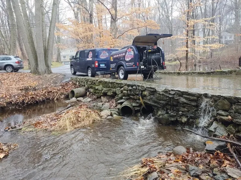 Work truck parked on a stone bridge over a flowing creek in a wooded area during fall.